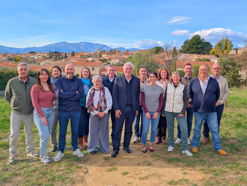 L'équipe de campagne de Michel Thiriet, le maire sortant, "Ensemble, agir pour Tresserre 2026 !" debout en deux rangs au Bois Pous à Tresserre. Le village est en arrière-plan : on apercoit le clocher de l'église St Saturnin de Tresserre et le Vallespir (Roc de Frausa, Roc Saint Sauveur...)

